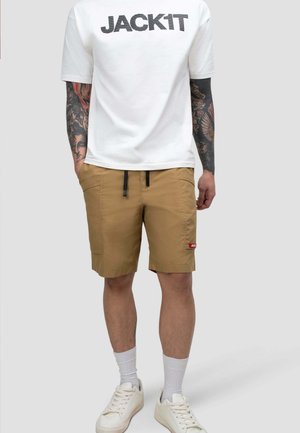Mężczyzna w białej koszulce z napisem „JACKIT”, beżowych szortach z przednimi kieszeniami, białych skarpetkach i białych tenisówkach, stoi z rękami w kieszeniach.
