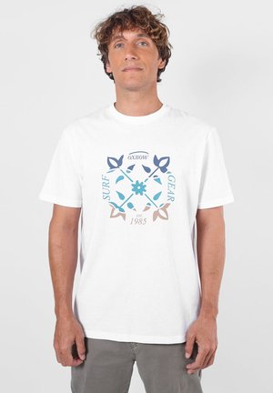 Homme aux cheveux bouclés portant un t-shirt blanc avec un motif floral bleu et marron et le texte "Oxbow Surf Gear est. 1985", debout devant un fond blanc.