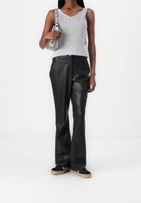 Top grigio a costine senza maniche, pantaloni a campana in ecopelle nera e sneakers nere con suole color nocciola. Modella che tiene una borsa a tracolla metallica.