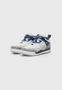 Weiße Sneakers mit marineblauen Akzenten, Klettverschlüssen, strukturiertem grauem Panel und gemusterter Sohle mit einem markanten schwarz-weißen Design.