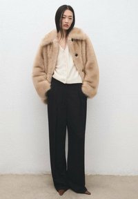 Chaqueta corta de piel sintética en tono marrón claro con botones negros, combinada con una rebeca crema y pantalones negros de talle alto. Silueta elegante, textura suave.