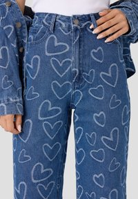 Vaqueros de mezclilla azules con un diseño de estampado de corazones, de cintura alta y con bolsillos delanteros. Material Texturizado con un aspecto desgastado. Cierre con botón metálico.