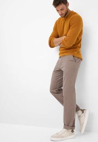 Un pull orange en maille et un chino gris clair porté avec des baskets beige. La tenue présente une coupe décontractée avec des poignets côtelés et des poches latérales.