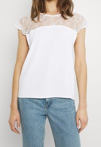 Weißes kurzärmliges Top mit einem Spitzenbestickten Schulterbereich und floralem Muster, geripptem Design und lockerer Passform, kombiniert mit hellblauen Jeans.