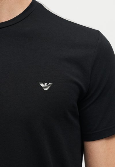 Μαύρο κοντομάνικο μπλουζάκι με στρογγυλή λαιμόκοψη και μικρό λευκό ριγέ λογότυπο αετού στο αριστερό στήθος, φορεμένο από άτομο.