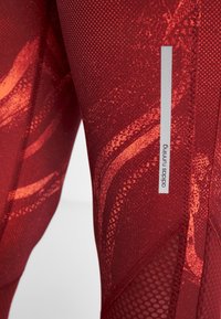 Czerwone legginsy do biegania z strukturalnym wzorem i pomarańczowymi akcentami, siateczkowymi detalami oraz srebrnym logo adidas z boku.