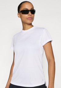 Weißes athletisches T-Shirt mit kurzen Ärmeln, rundem Halsausschnitt und glatter Textur, kombiniert mit dunkler Sonnenbrille. Dezentes Logodetail am Saum.