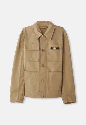 Tan jacket made of cotton, featuring a pointed collar, four patch pockets, gold buttons, and a detail with a black accent on the chest.