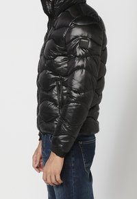 Persona che indossa una giacca a vento nera lucida con una toppa con il logo Koroshi sulla manica e jeans blu, in piedi di lato contro uno sfondo uniforme.