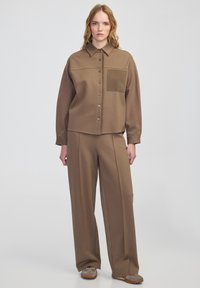 Brązowa koszula z guzikami i kieszenią na piersi, w zestawie z szerokimi brązowymi spodniami. Gładki materiał; luźny krój; dopasowane buty z teksturowanymi akcentami.