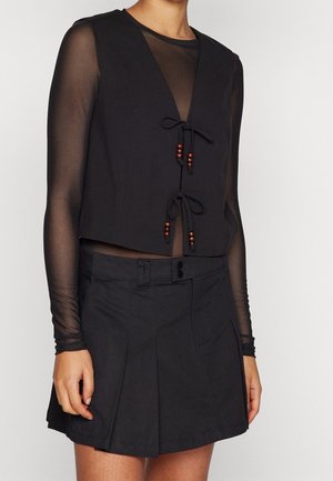Woman wearing black sleeveless vest with wooden bead ties over sheer long-sleeve top and black pleated mini skirt.