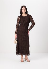 Braunes Spitzenkleid mit langen Ärmeln und floralem Muster, knielanger Saum. Das Model hält eine kleine beigen Handtasche und trägt silberne Absatzsandalen.