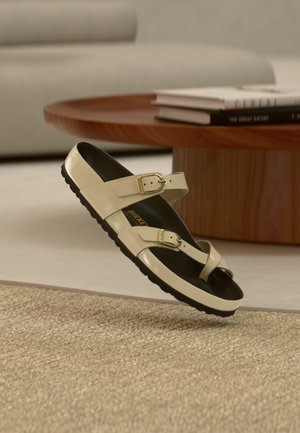 Sandale beige avec deux brides à boucle dorée et semelle noire flottant au-dessus d'un tapis beige tissé, table basse en bois et livres en arrière-plan.