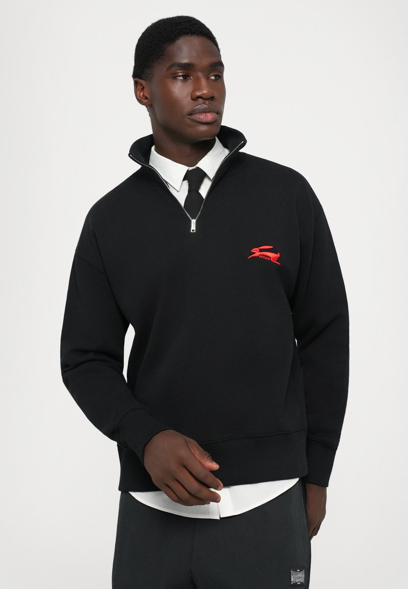 Man in een zwart quarter-zip sweater met een rood konijnenlogo, een wit shirt, een zwarte stropdas en donkere broek tegen een effen achtergrond.