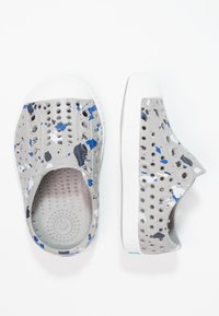 Zapatos grises sin cordones hechos de material perforado, con un patrón de salpicaduras multicolor en azul y blanco, y una suela de goma blanca.