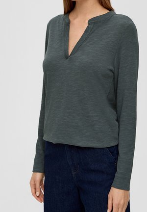 Kvinde iført en mørkegrøn langærmet trøje med en V-udskæring og mørkeblå jeans, stående mod en ensfarvet baggrund.