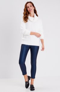 Witte, fluffy gebreide sweater met een col, gecombineerd met glanzende donkerblauwe skinny jeans en zwarte loafers, op een model tegen een neutrale achtergrond.