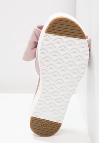 Semelle d'une chaussure UGG rose clair avec une bande de roulement blanche texturée et des grips en caoutchouc marron au talon et à l'avant.