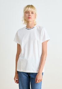 Witte katoenen t-shirt met korte mouwen en een ronde hals, met een zwarte logo-print op de kraag. Eenvoudig en onopgesmukt ontwerp.