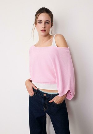 Giovane donna con capelli raccolti che indossa un maglione rosa largo con spalle scoperte sopra una canottiera bianca e jeans blu scuro, con le mani nelle tasche.