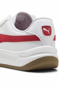 Sneakers bianchi con una striscia rossa e suola beige, dotati di un colletto imbottito e dettagli testurizzati. Logo impresso sul tallone.