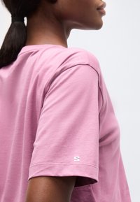 Άτομο που φοράει ροζ μαλακό πουκάμισο με κοντά μανίκια και μικρό λευκό λογότυπο "S" στο μανίκι, εμφανιζόμενο από το πλάι και πίσω.