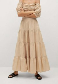 Vaaleanbeige kerroksellinen maksimekko, jossa on olkapäiltä paljastavat hihat. Mekossa on rypytetty yläosa ja laskeutuva siluetti. Yhdistetty mustiin sandaaleihin.