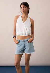 Bela halter crop majica z zavitim dizajnom, v kombinaciji z svetlo modrimi denim kratkimi hlačami z raztrganimi robovi in subtilnimi okraski.