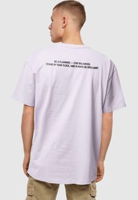 Lichtpaarse t-shirt met korte mouwen en een ontspannen pasvorm. De tekst op de achterkant in zwart leest: "WEES EEN FLAMINGO - BLIJF IN BALANS..."