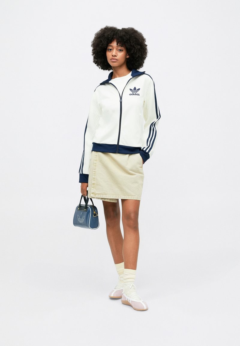 White zip-up jacket with navy accents and three striped sleeves, paired with a beige skirt. Accessory: small blue handbag with logo.