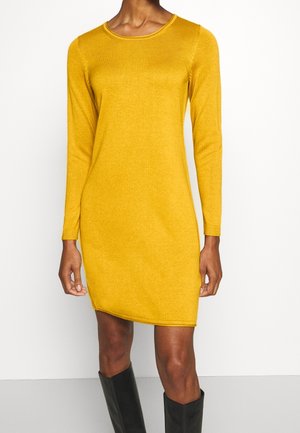 Kobieta w musztardowo-żółtej długiej dzianinowej sukience z długim rękawem, w czarnych czółenkach za kolano, stojąca na jednolitym tle.