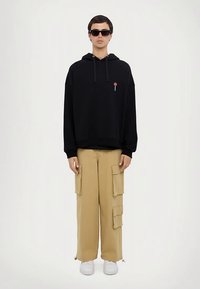 Felpa nera con cappuccio, tasca anteriore e logo, abbinata a pantaloni cargo beige con molteplici tasche. Sneakers bianche completano il look.