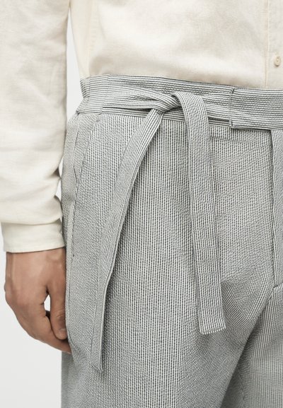 Άνδρας με ανοιχτό μπεζ πουκάμισο και γκρι παντελόνι με ρίγες και υφασμάτινη ζώνη δεμένη σε κόμπο στη μέση, χέρι χαλαρωμένο δίπλα στο σώμα.