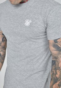 Grijze T-shirt met korte mouwen, gemaakt van een zachte, textuurstof, met een wit geborduurd logo op de linkerkant van de borst.