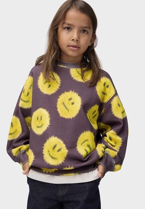 Kind mit langen braunen Haaren, das einen lila Pullover mit gelben, flauschigen Smiley-Gesichtern und dunkelblauen Jeans trägt, Hände in den Taschen.