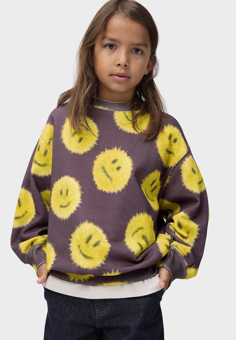 Enfant aux longs cheveux bruns portant un sweat violet avec des visages souriants jaunes duveteux et un jean bleu foncé, mains dans les poches.