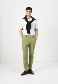 Camiseta blanca, pantalones verdes, suéter azul marino drapeado alrededor del cuello, zapatos negros. La persona está de pie frente a un fondo neutro.