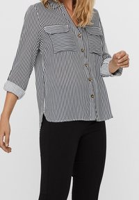 Camicia a maniche lunghe a righe in bianco e nero, con due tasche sul petto, maniche arrotolate e orlo curvo. Tessuto leggero e liscio.