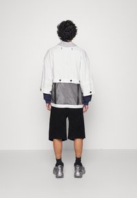 Biała, oversize'owa kurtka z siateczkowym tyłem, zestawiona z czarnymi szortami w wzory i przezroczystymi sneakersami. Posiada szerokie rękawy i guziki.