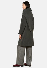 Cappotto grigio in lana con colletto drappeggiato, abbinato a pantaloni larghi lucidi e scarpe bordeaux. Il tessuto ha una texture liscia e un design a strati.