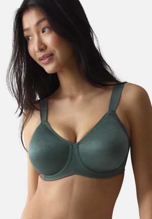 Femme aux longs cheveux foncés portant un soutien-gorge vert foncé avec des bretelles larges, regardant légèrement sur le côté sur un fond uni.