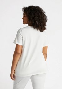 Top blanco de manga corta con puños enrollados, fabricado en una tela suave, con una silueta relajada y aberturas laterales. La vista trasera revela un diseño suave y minimalista.