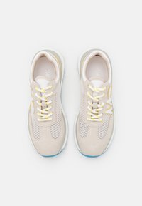 Baskets beige clair avec un dessus en maille, des accents en daim, des lacets jaunes et une semelle blanche texturée avec un détail bleu.