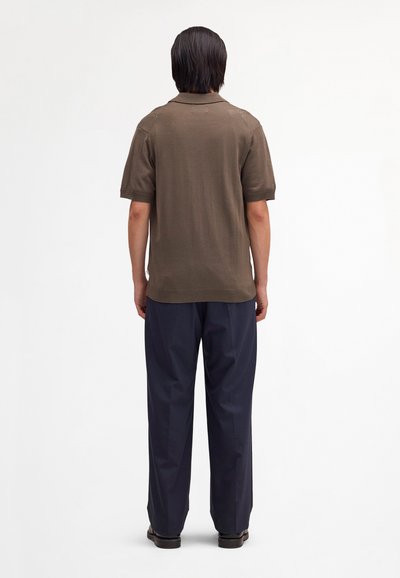 Homme aux cheveux noirs raides, vu de dos, portant un polo marron à manches courtes, un pantalon large bleu marine et des chaussures noires sur un fond uni.