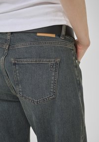 Tumši zili džinsi ar klasisku griezumu, ar aizmugurējo kabatu ar šuvju detaļām un brūnu ādas plāksteri virs jostas līnijas.