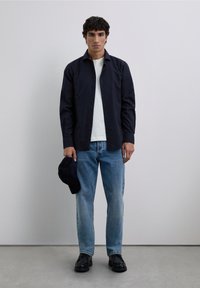 Hombre de pie frente a un fondo liso, vistiendo una camisa marina sobre una camiseta blanca, jeans azules, zapatos negros y sosteniendo una gorra marina en la mano izquierda.