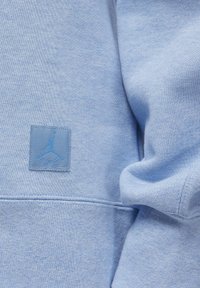 Sudadera azul claro con un tejido texturizado, que presenta un parche del logo Jumpman azul cosido en el lado izquierdo del pecho, con costuras y puntadas visibles.