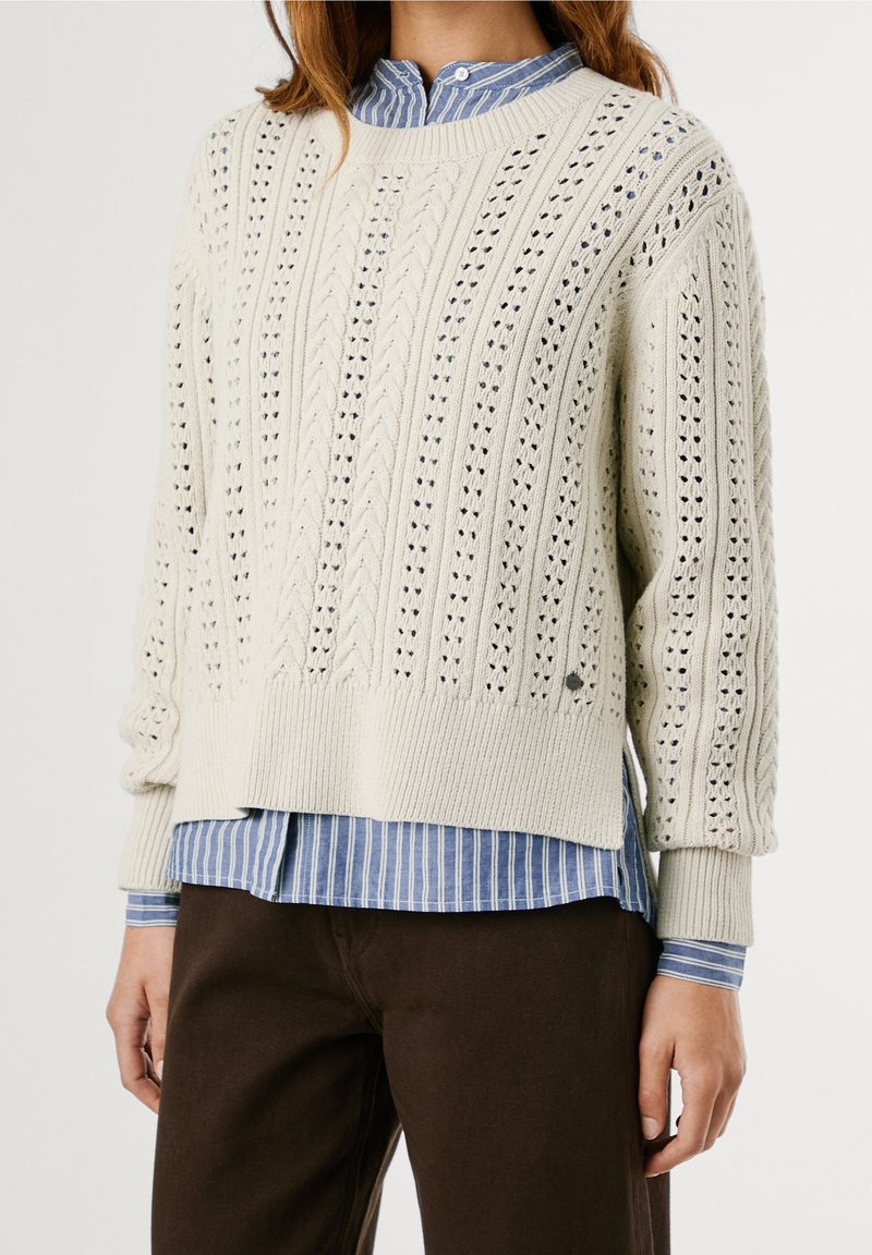 Femme portant un pull crème à torsades par-dessus une chemise à rayures bleues et des pantalons marron foncé, debout devant un fond blanc.