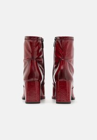 Bottines en cuir verni rouge avec un talon carré, présentant une fermeture à glissière à l'arrière et une texture lisse et brillante.