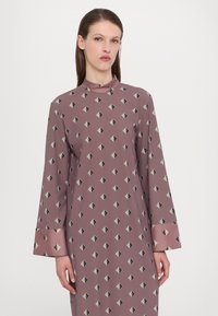 Langärmliges Kleid aus mauve-farbenem Stoff mit einem geometrischen Muster aus Diamanten in Schwarz und Hellgrün. Hoher Kragen und umschlagbare Ärmel.